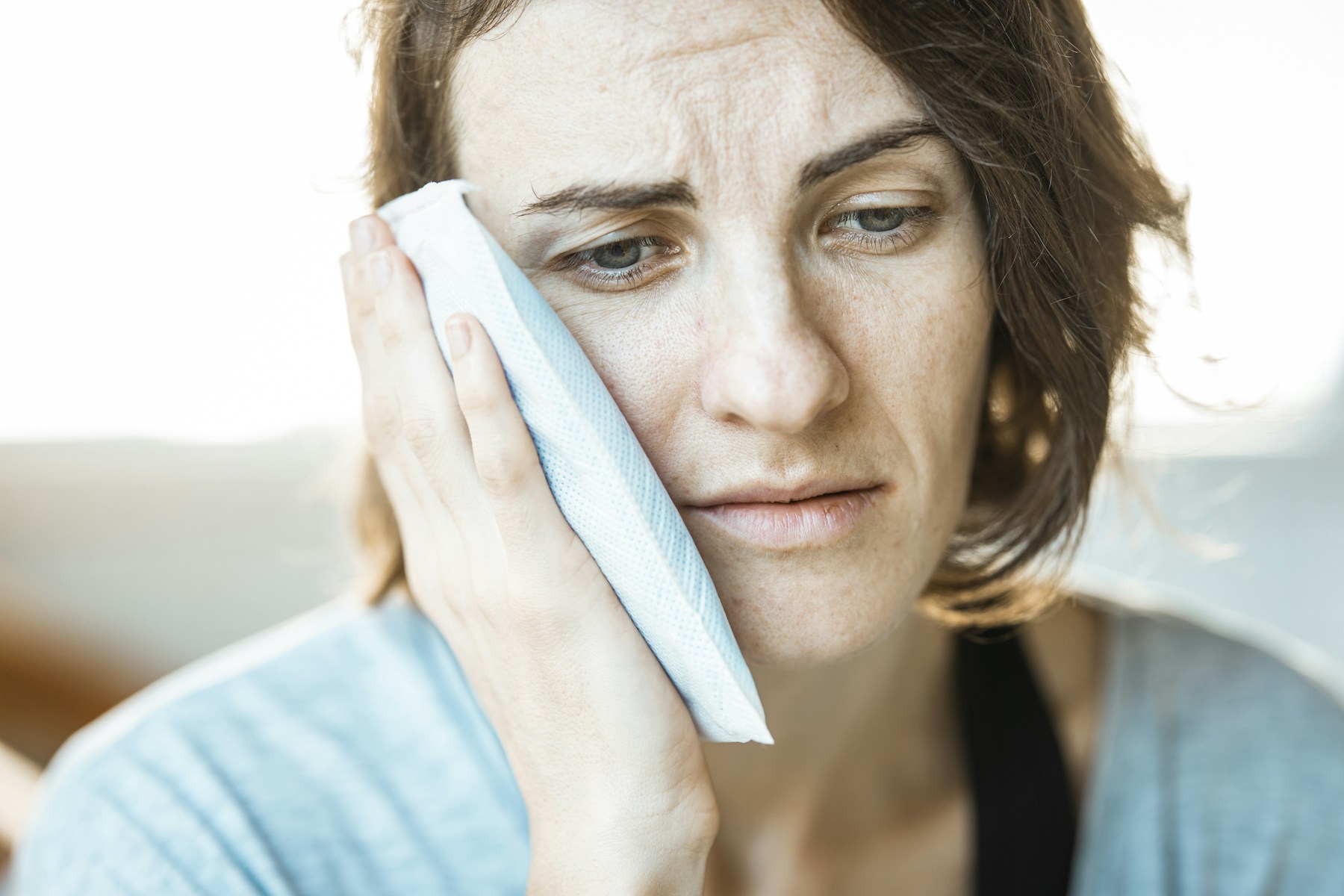 A woman looking down while holding a cold compress against her aching cheek.