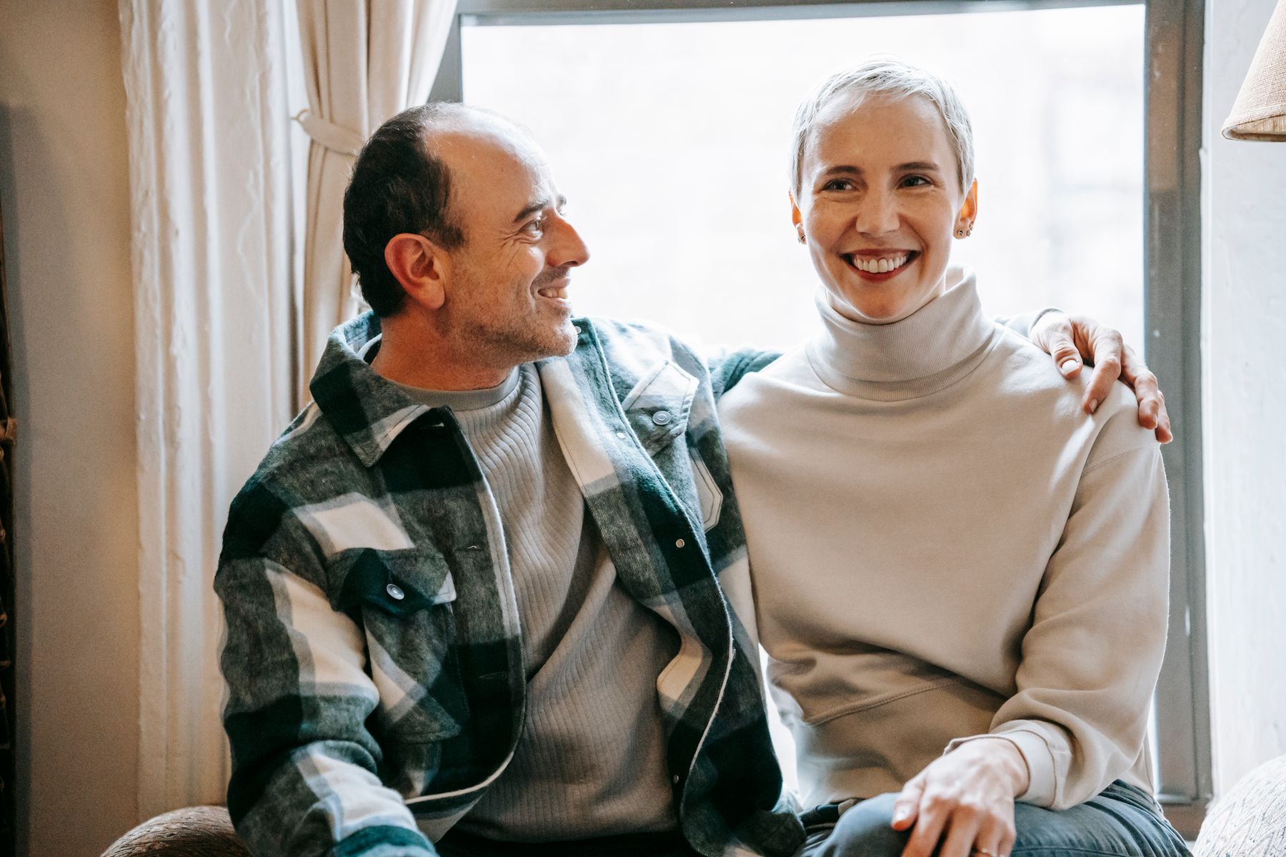 A mature couple smiling together beside a window.