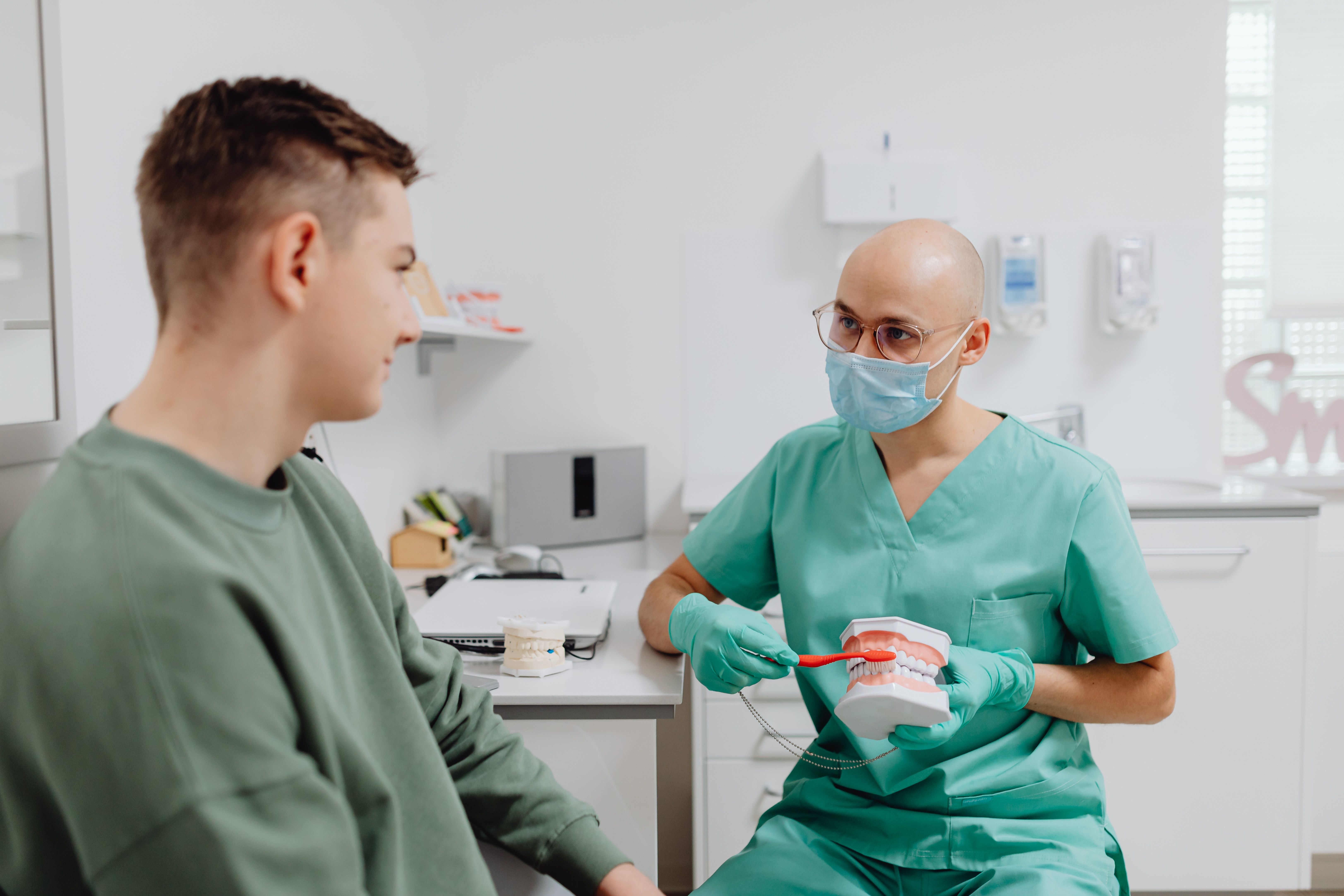 A male dentist explains brushing with a dental model to a patient in a green sweatshirt, whose face is visible in profile.